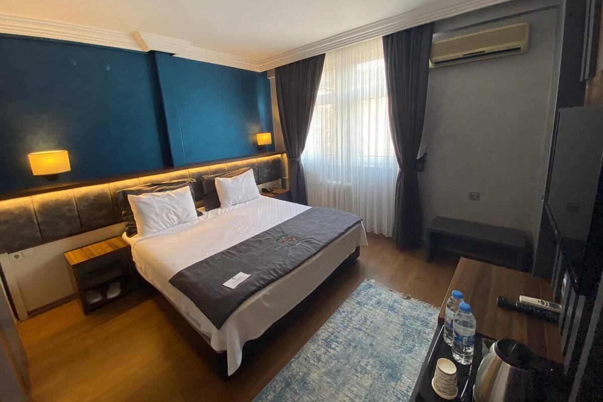 A hotel room with a large bed, a bedside table, a lamp, a TV, and a window with curtains.