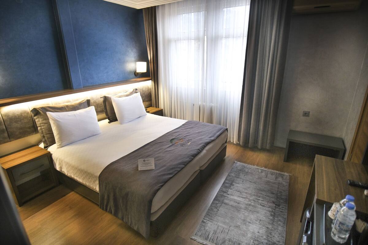 A hotel room with a bed, bedside tables, a nightstand, a desk, and a window with curtains.