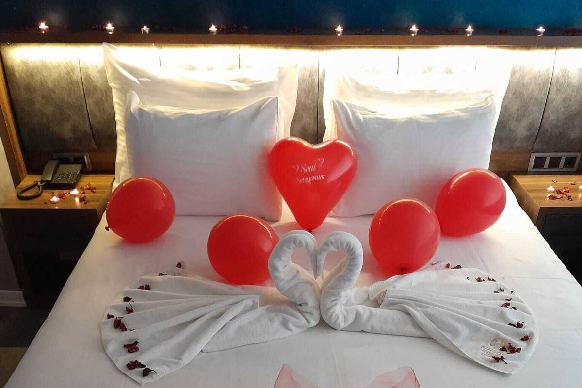A bed with white linens, red heart-shaped balloons, and a red bow.