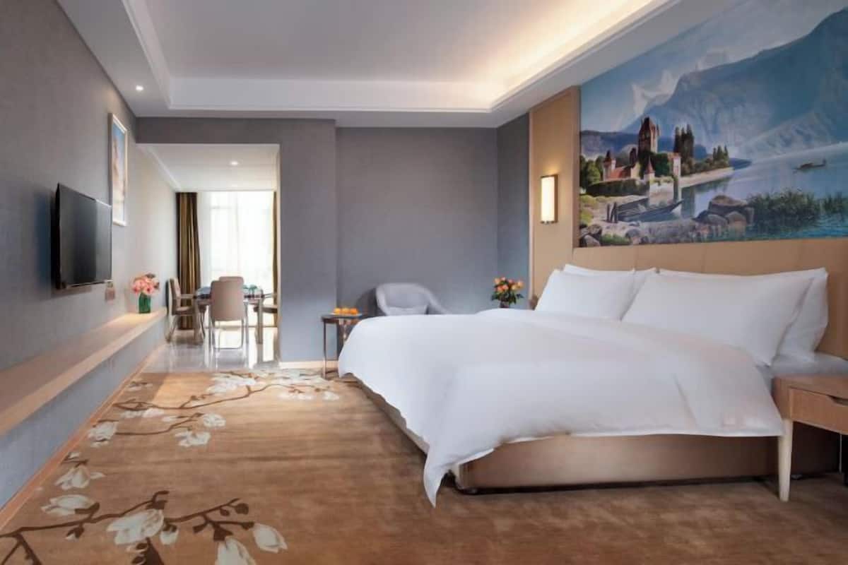Una habitación de hotel con una cama grande, un televisor de pantalla plana en la pared, un cuadro en la pared y piso alfombrado.