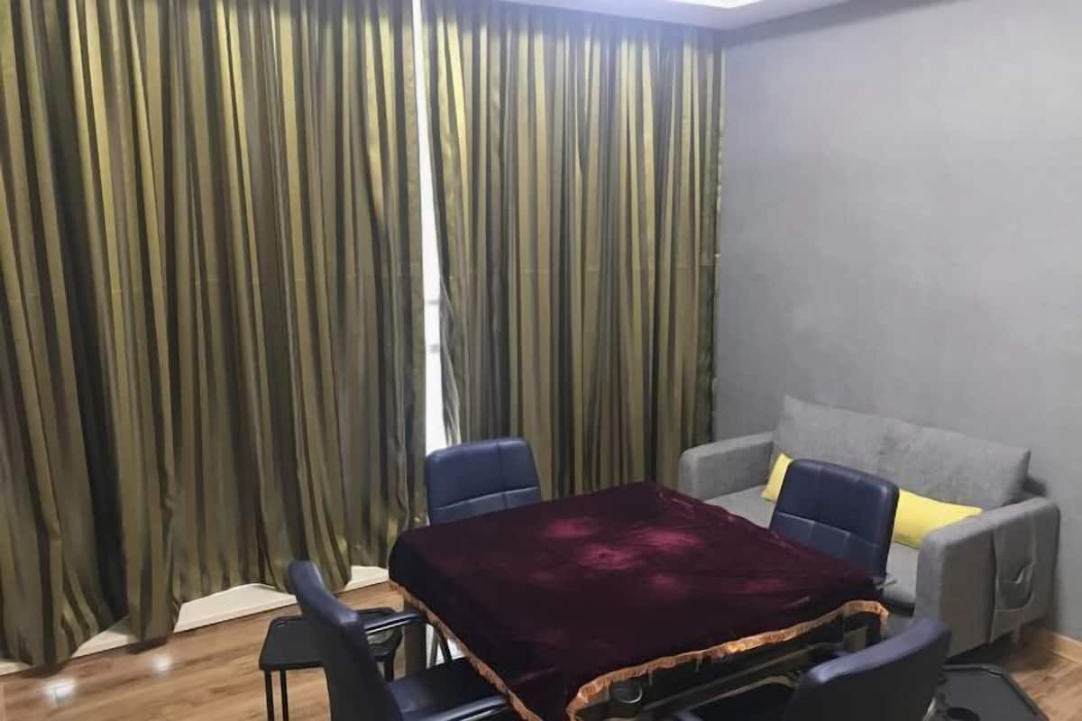 Una habitación con una mesa cubierta por un paño oscuro, dos sillas, un sofá con cojines y una cortina de rayas verticales.