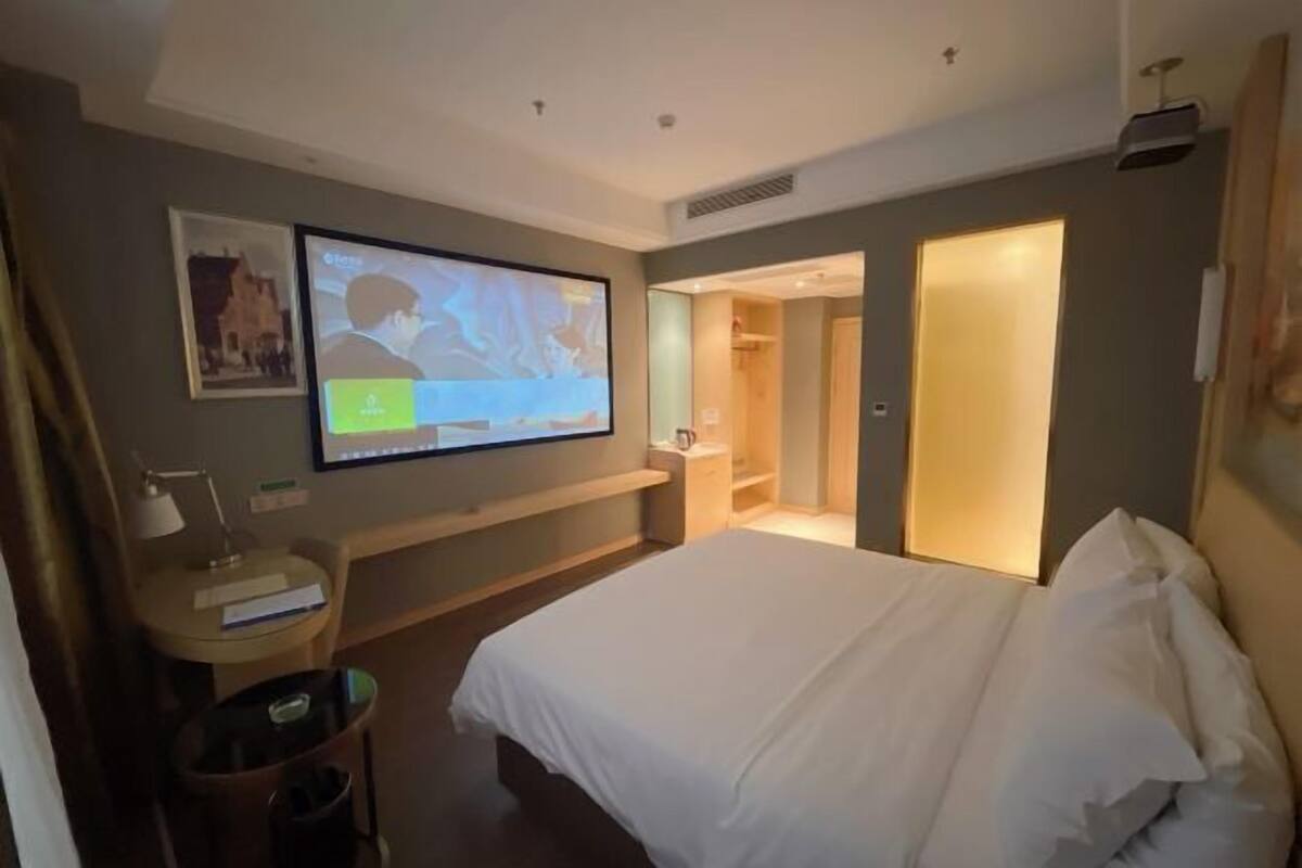 Habitación de hotel con cama, televisor montado en la pared, escritorio con lámpara y un pequeño cubo de basura.