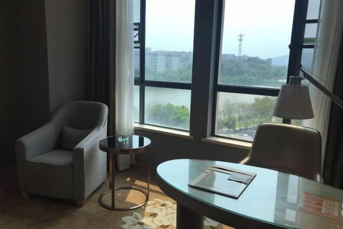 A hotel room with a glass table, two chairs, a lamp, and a view of buildings and greenery through large windows.