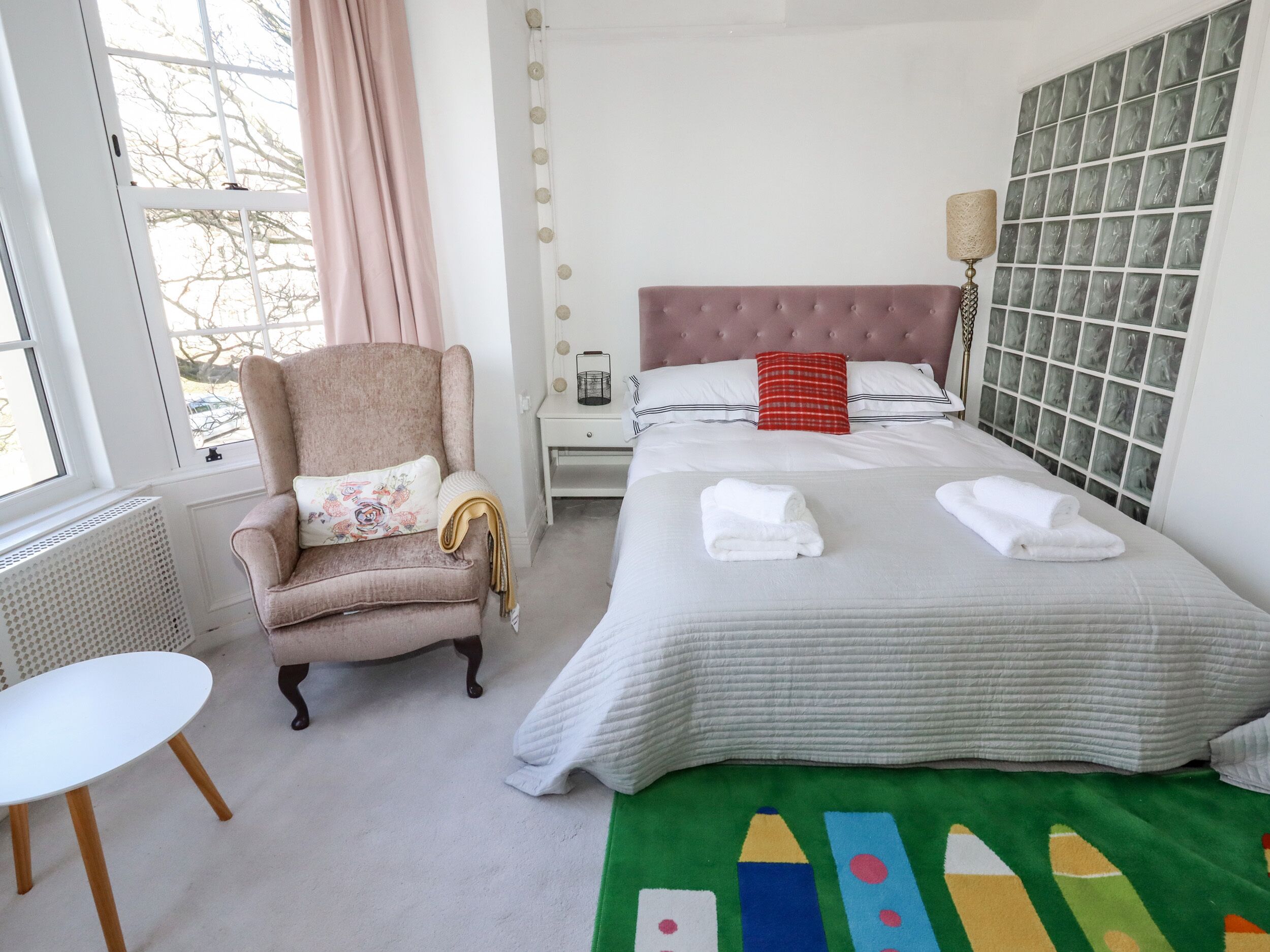 Een keurig opgemaakt slaapkamer met een bed, een stoel, een klein tafeltje en een raam met gordijnen.