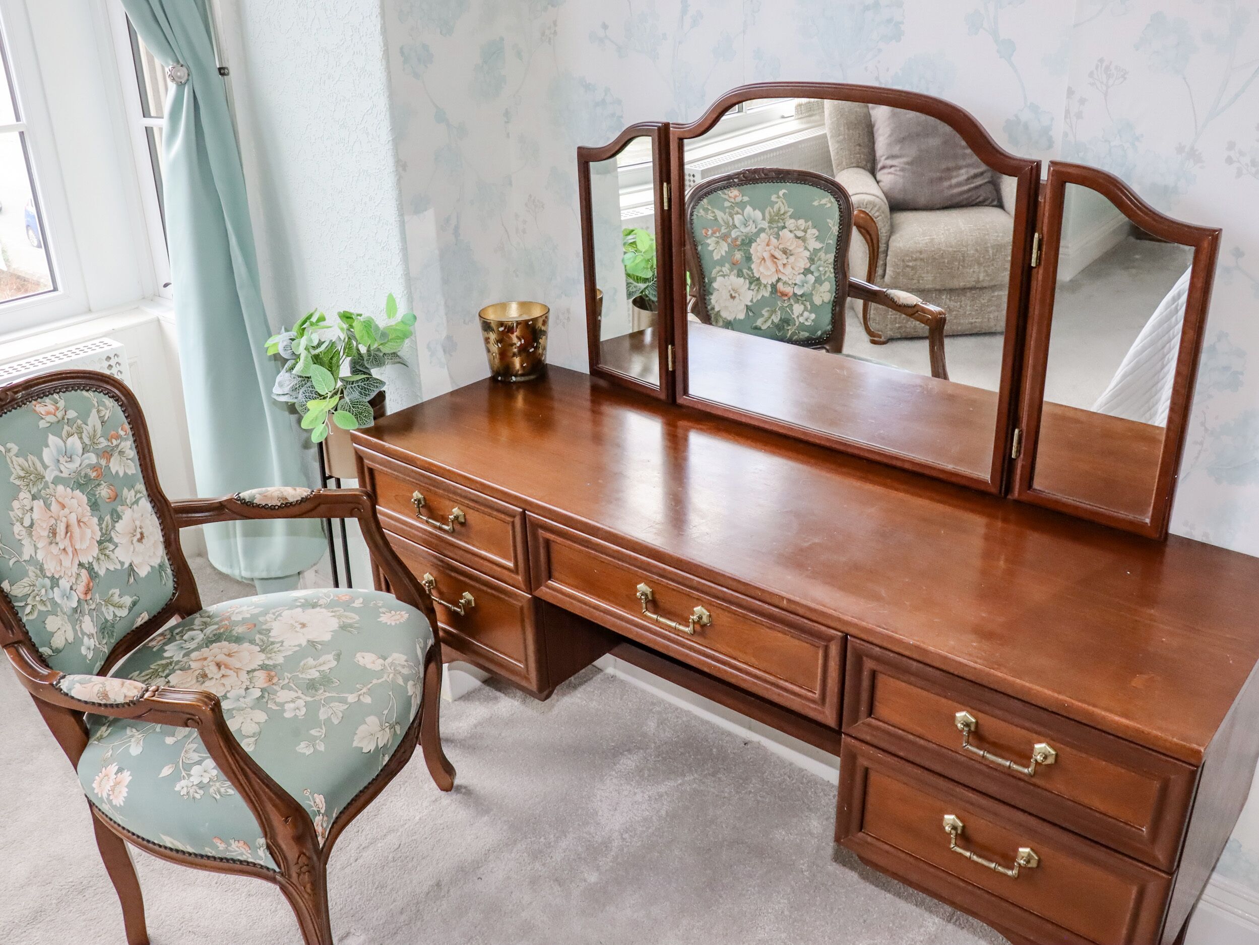 A wooden dresser with drawers and a mirror, a floral upholstered chair, and a potted plant.
