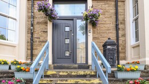 Un edificio con una porta grigio scuro, una piccola finestra e il numero '112' sopra la porta. Ci sono vasi fioriti con diversi fiori sui gradini che conducono alla porta.