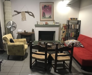 A living room with a fireplace, a red sofa, a wooden dining table with chairs, a bookshelf, and a wall-mounted fan.