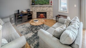 Een woonkamer met een open haard, grijze banken, een salontafel en een televisie bevestigd aan een stenen muur.