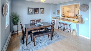Een eetruimte met een houten tafel en stoelen, een bank, een vloerkleed, en een keuken die zichtbaar is door een open doorgang.