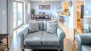 Een woonkamer met een grijze bank, een eettafel en een keuken die zichtbaar is door een open deur.