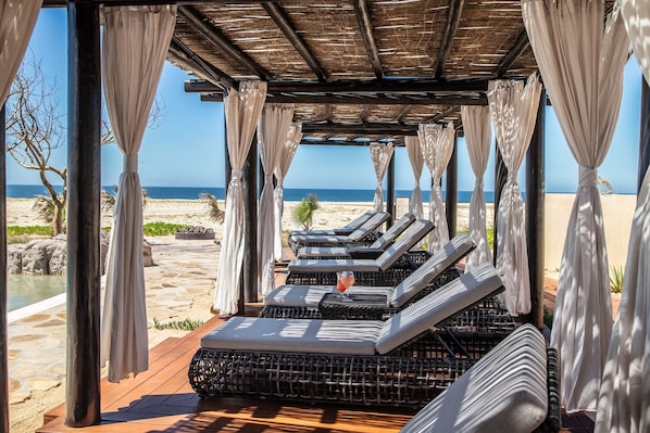 A beachside lounge area with cushioned loungers, a wooden deck, and a view of the ocean.