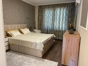 Un dormitorio con una cama grande, una mesita de noche, una pequeña mesa de madera y una ventana con cortinas.