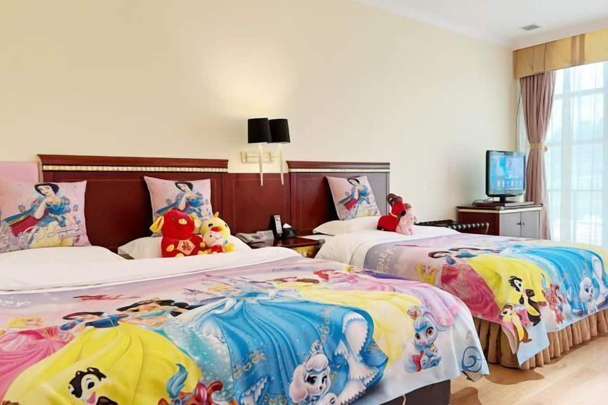 A hotel room with two beds, each adorned with Disney-themed bedding. There is a television on a stand to the right, and a nightstand with a lamp and phone.