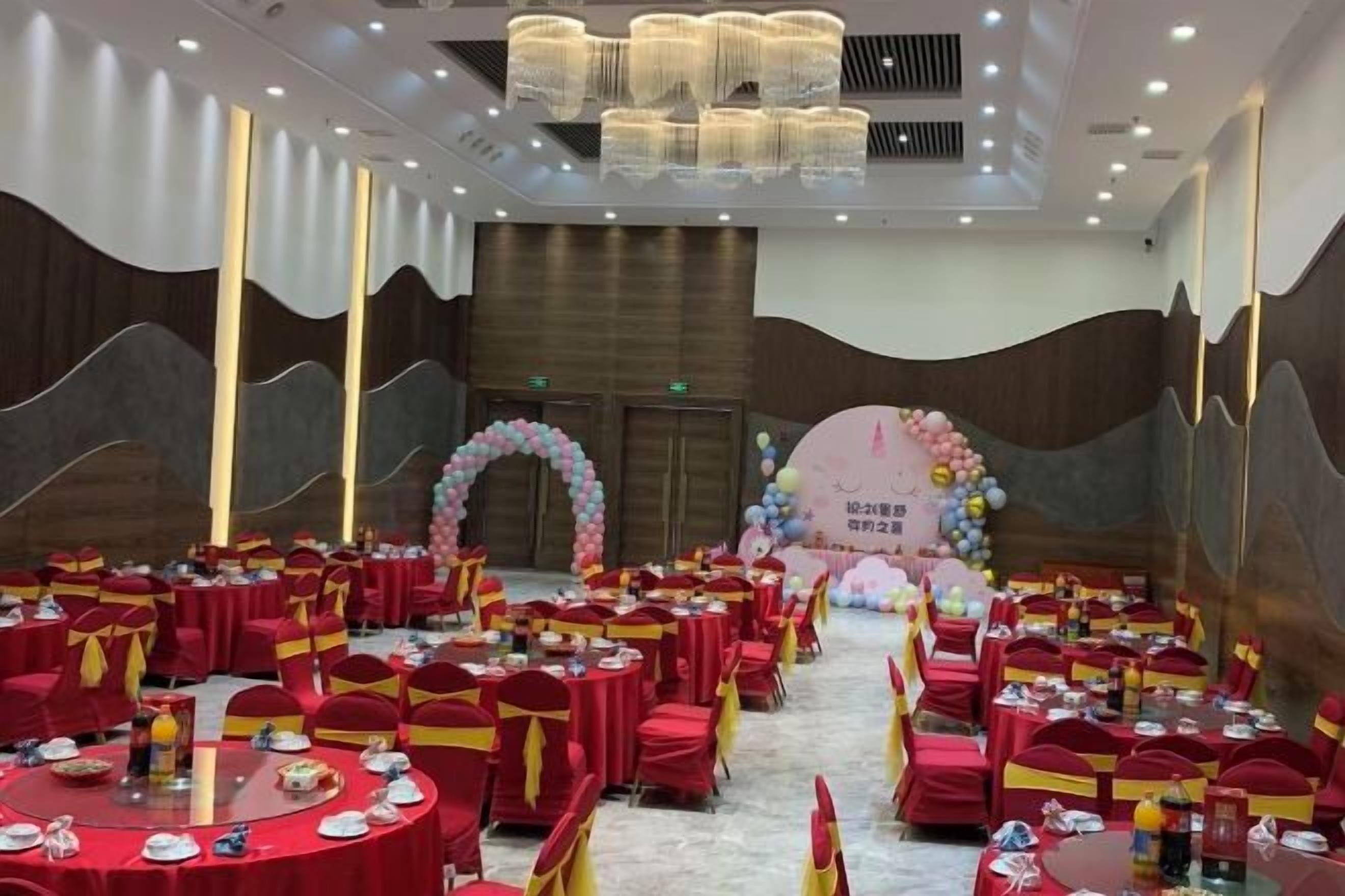 Un salón de banquetes con mesas redondas, sillas rojas y un escenario con un fondo floral.