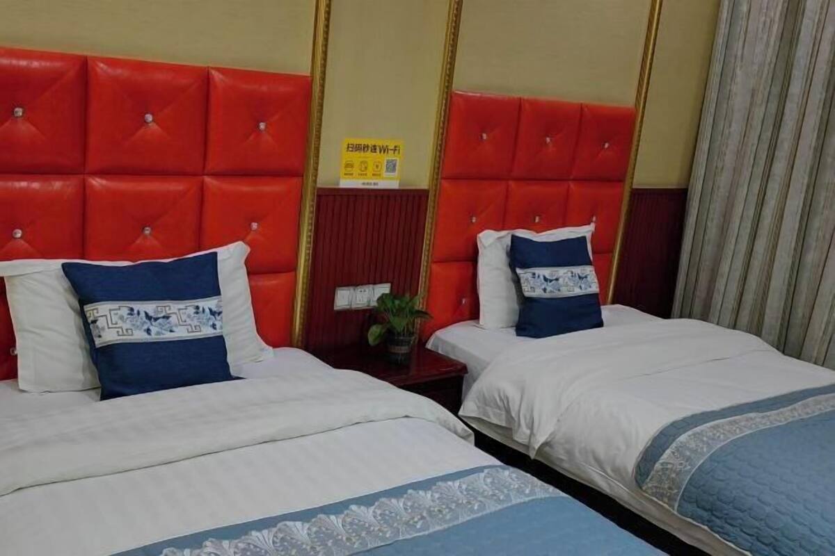 A hotel room with two beds, each with white bedding and blue decorative pillows. The headboards are red with a tufted design. There is a small table with a plant and a sign on the wall.