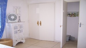 Una habitación con un armario blanco, un ventilador y un taburete de mimbre.