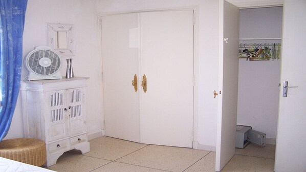 Ein Zimmer mit einem weißen Kleiderschrank, einem Ventilator und einem Korbstuhl.
