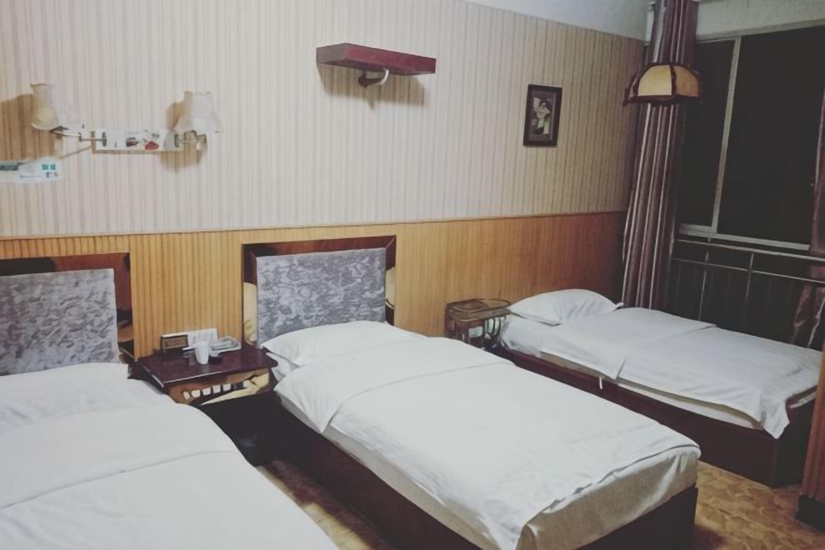 Ein Hotelzimmer mit drei Einzelbetten, einem Holzkopfstück, einem kleinen Tisch mit Lampe und einem Fenster mit Vorhängen.
