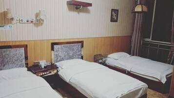Ein Hotelzimmer mit drei Einzelbetten, einem Holzkopfstück, einem kleinen Tisch mit Lampe und einem Fenster mit Vorhängen.
