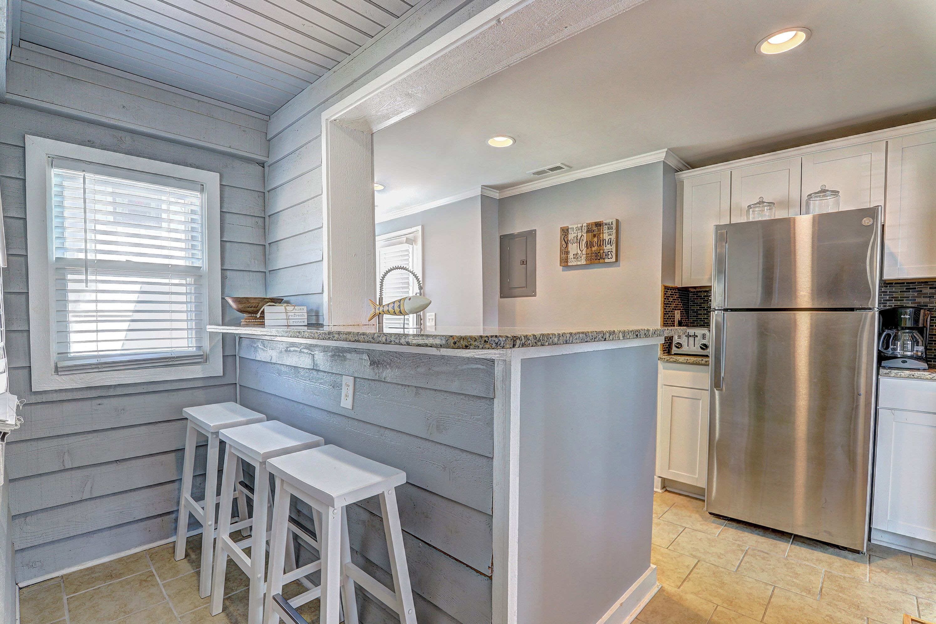 A kitchen with a central island, white bar stools, a stainless steel refrigerator, and a window with blinds.