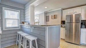 A kitchen with a central island, white bar stools, a stainless steel refrigerator, and a window with blinds. - Cottage on the Rocks - 3 Bedrooms - Walk to Beach - Outdoor Seating - Pet Friendly (North Myrtle Beach)