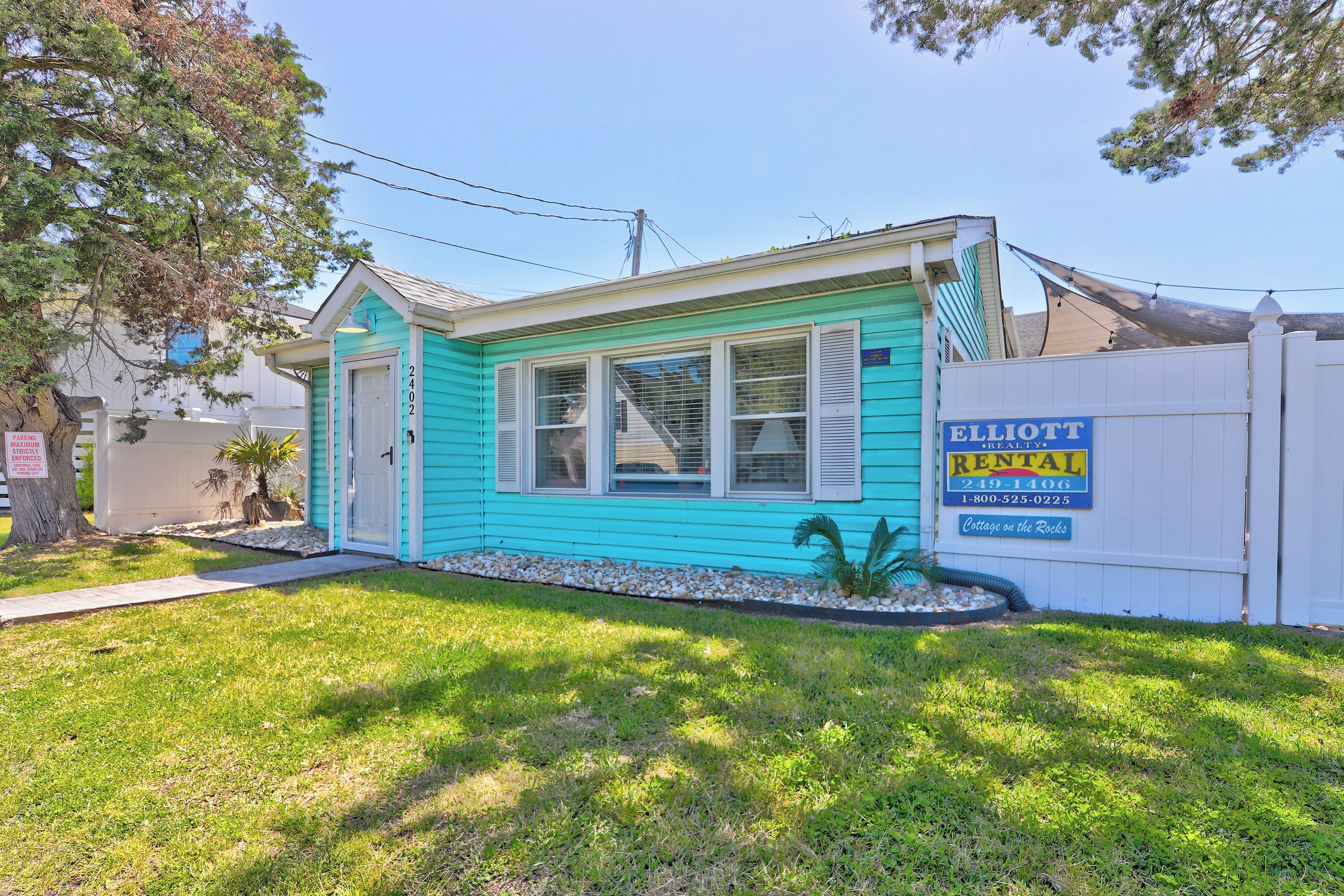 A turquoise rental cottage with a fenced yard, a sign advertising rental, and a small garden area.