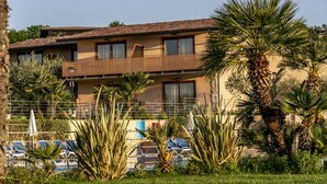 A two-story building with a balcony, surrounded by palm trees and a pool area with lounge chairs. - Apartment in Manerba with barbecue (Manerba sul Garda)