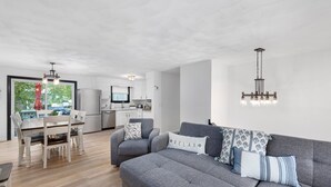 Ein modernes Wohnzimmer mit einer grauen Couch, einem Esstisch mit Stühlen und einer Küche im Hintergrund.