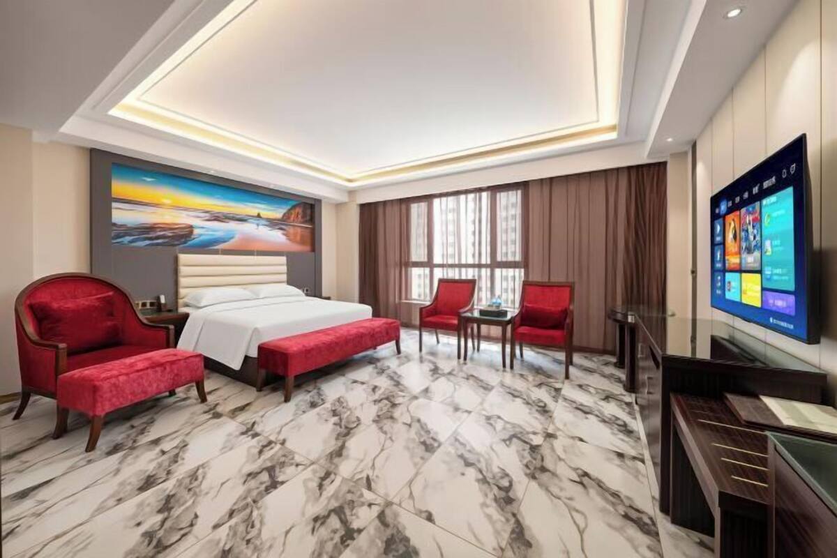 A hotel room with a bed, red armchairs, a TV, and a large window.