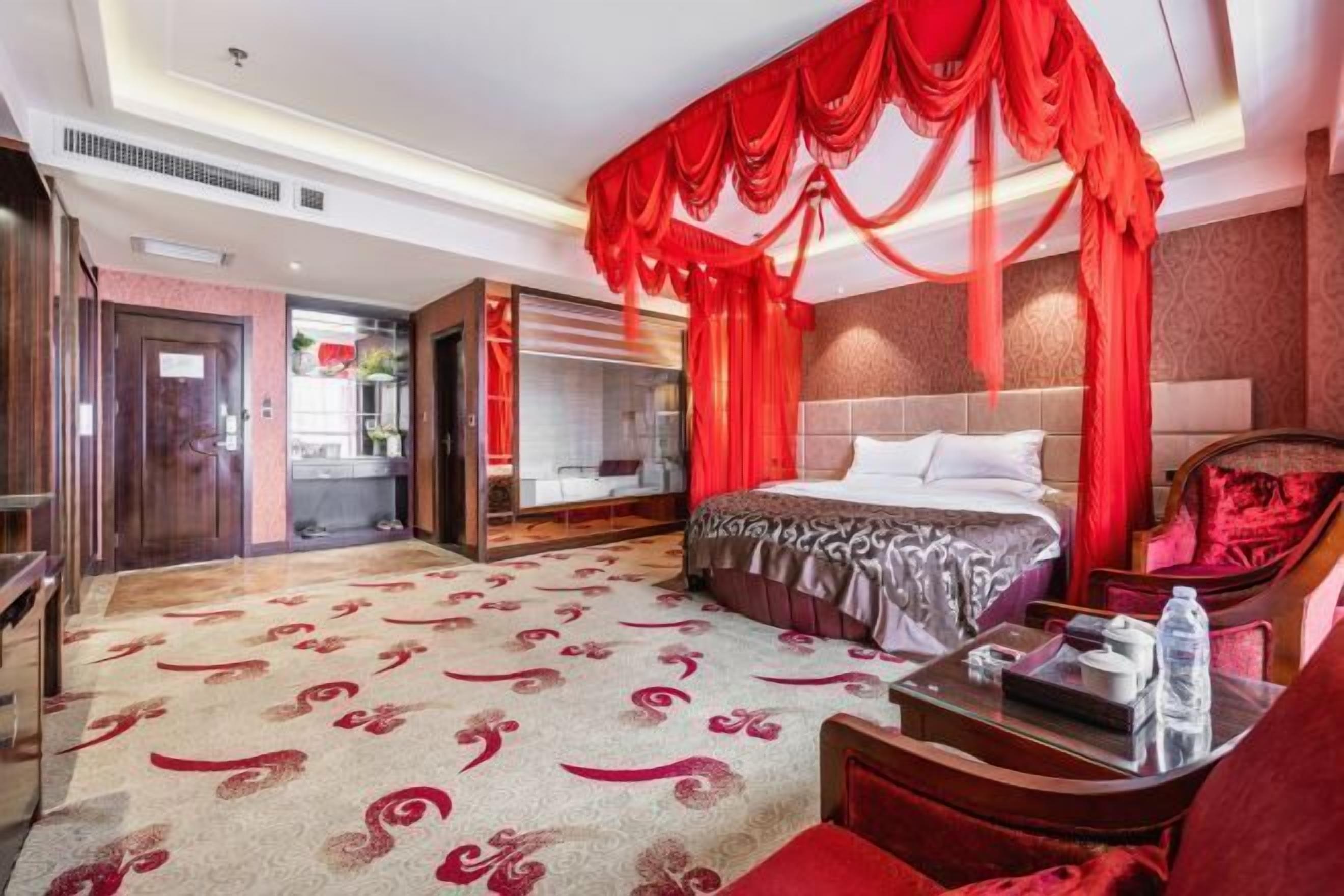 Habitación de hotel con una cama grande, cortinas rojas, cabecera de madera, un sofá rojo y una mesa de centro de vidrio.