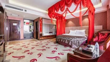 Habitación de hotel con una cama grande, cortinas rojas, cabecera de madera, un sofá rojo y una mesa de centro de vidrio.