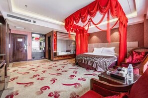 Habitación de hotel con una cama grande, cortinas rojas, cabecera de madera, un sofá rojo y una mesa de centro de vidrio.