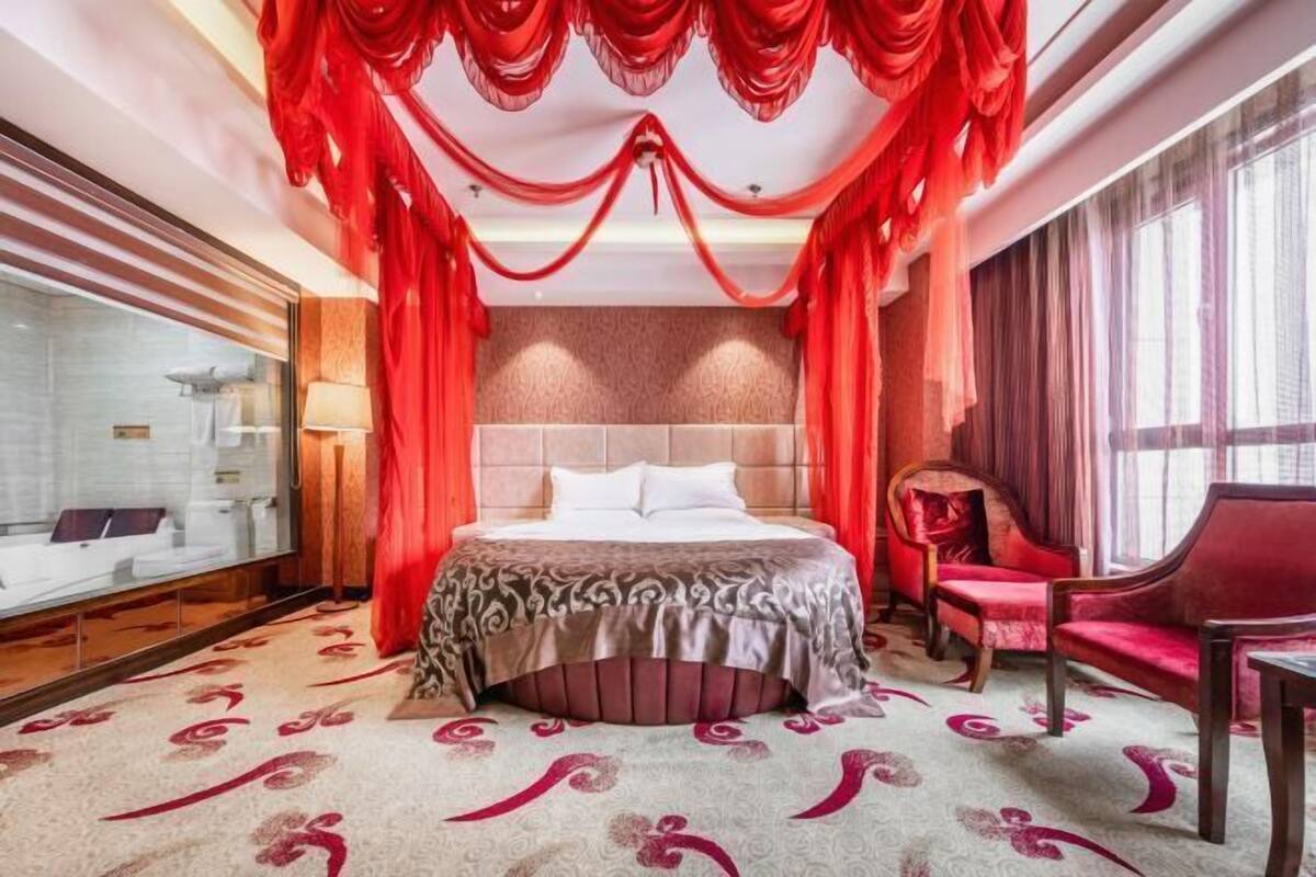 A luxurious hotel room with a large bed, a sitting area with a red chair, and a glass-enclosed bathroom.