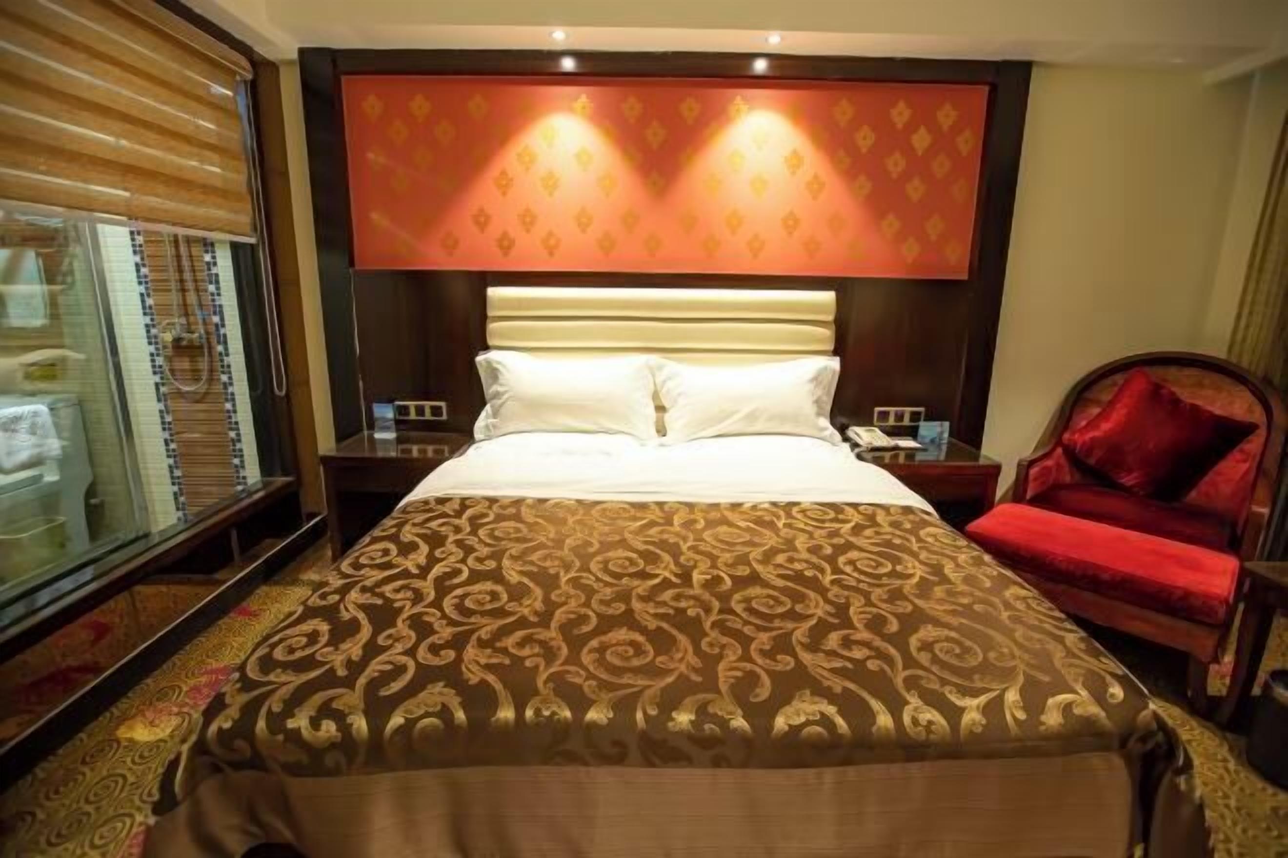 A hotel room with a large bed, a red chair, a glass shower enclosure, and a patterned headboard.