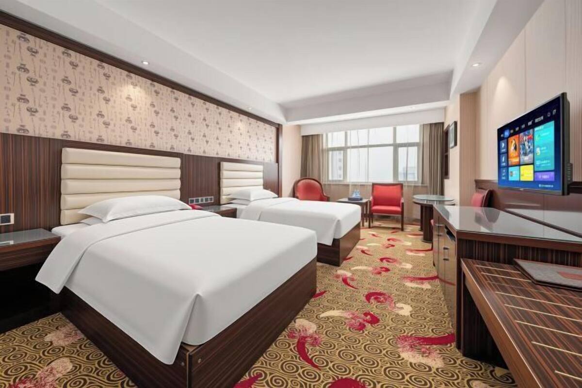 A hotel room with two beds, a desk, a chair, a TV, and a patterned carpet.