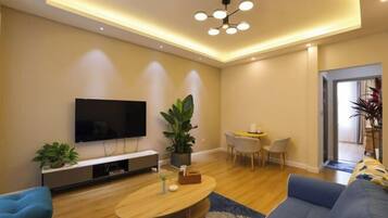 A modern living room with a flat-screen TV, a wooden coffee table, a blue sofa, and a dining area with a small table and chairs.