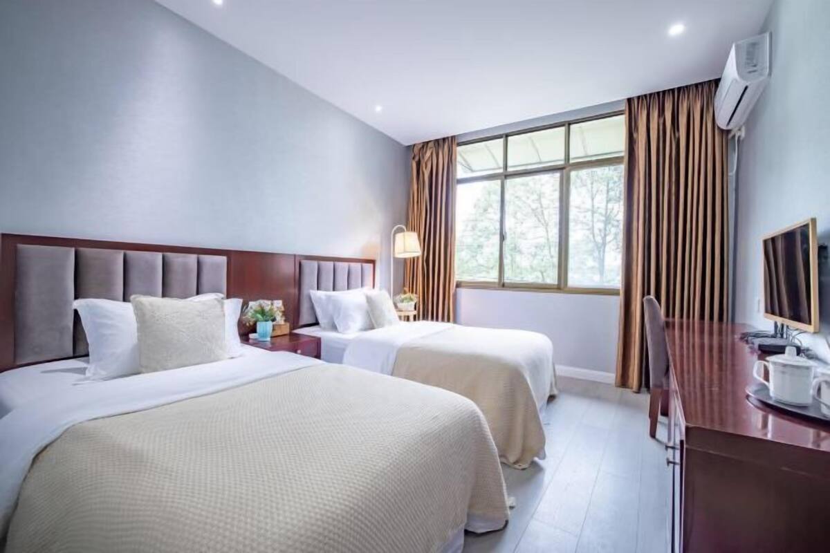 Hotellihuone, jossa on kaksi sänkyä, työpöytä, tuoli, lamppu ja suuri ikkuna, jossa on verhot.