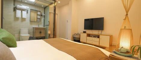 Habitación de hotel moderna con una cama grande, un televisor, una mesita de noche con lámpara y vistas al baño a través de una puerta de vidrio.