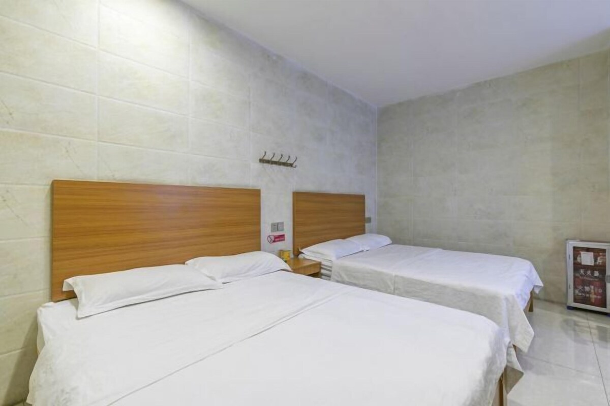 A hotel room with two single beds, wooden headboards, a small bedside table with a lamp, and a wall-mounted hook.