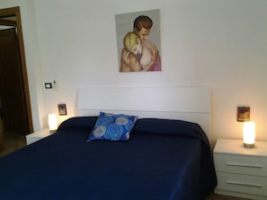 Een slaapkamer met een bed, een hoofdbord, een blauw kussen, nachtkastjes en een kunstwerk aan de muur.