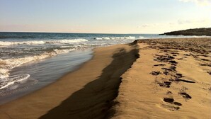 Ein Sandstrand mit Fußspuren, die zum Wasser führen, ein klarer Himmel und in der Ferne felsige Formationen.