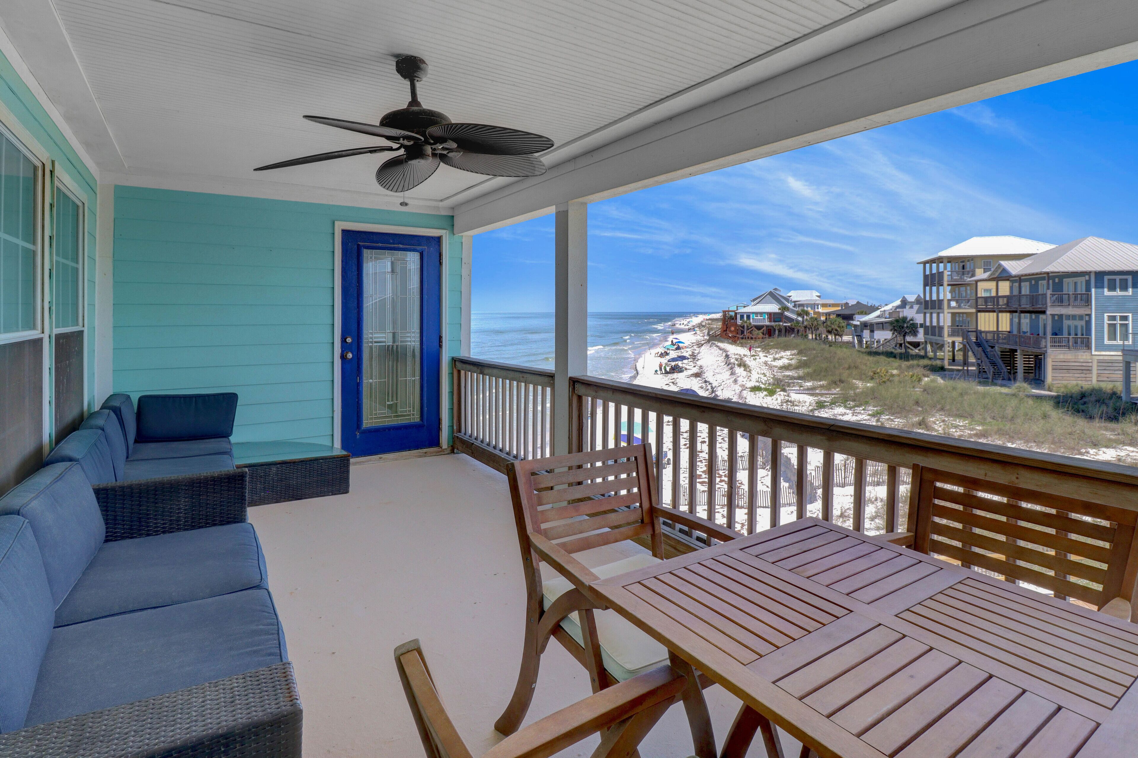 A covered porch with a view of the beach, a blue door, and a table with chairs.