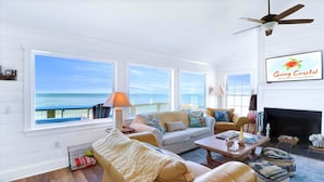 A spacious living room with large windows, a fireplace, and a ceiling fan. - 4 Bed/3 Bath North Cape San Blas Beach Front Home - Steps to the Water!! (Port St. Joe)