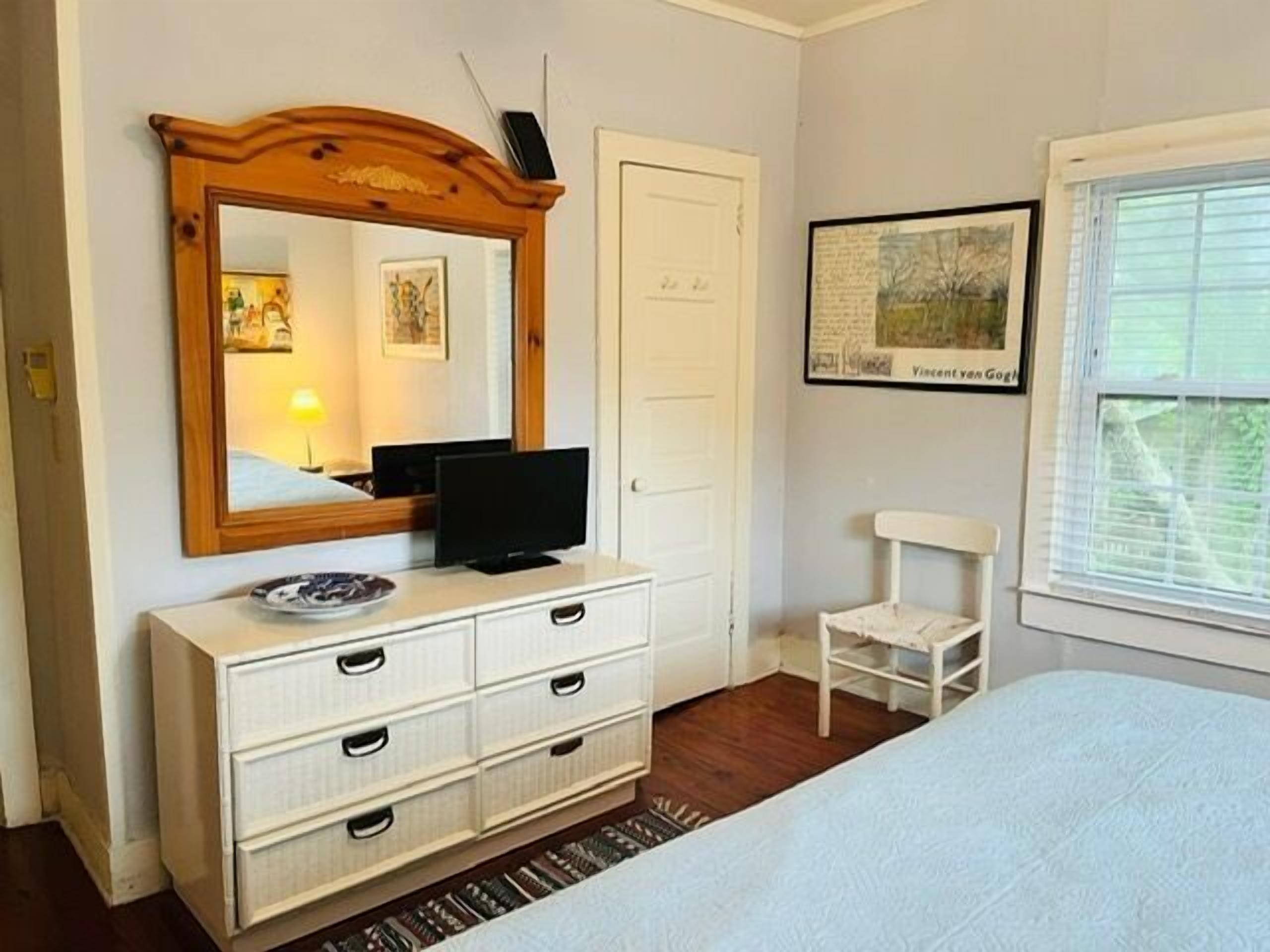 Una camera da letto con un letto, una cassettiera, una televisione, una sedia, un quadro appeso al muro e una finestra.
