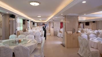 A banquet hall with round tables covered in white tablecloths and chairs, a central serving station, and a long corridor.
