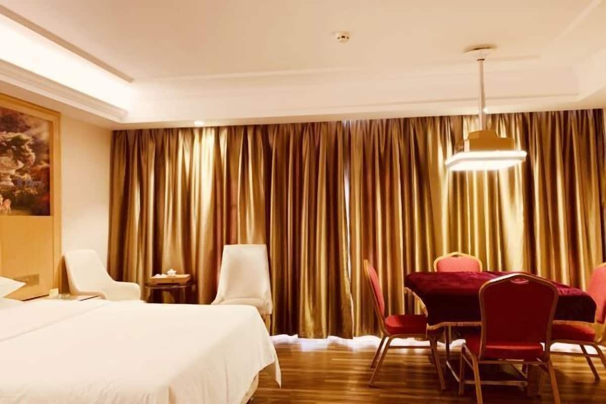 Quarto de hotel com cama, mesa de jantar com cadeiras, um quadro na parede e uma cortina grande.