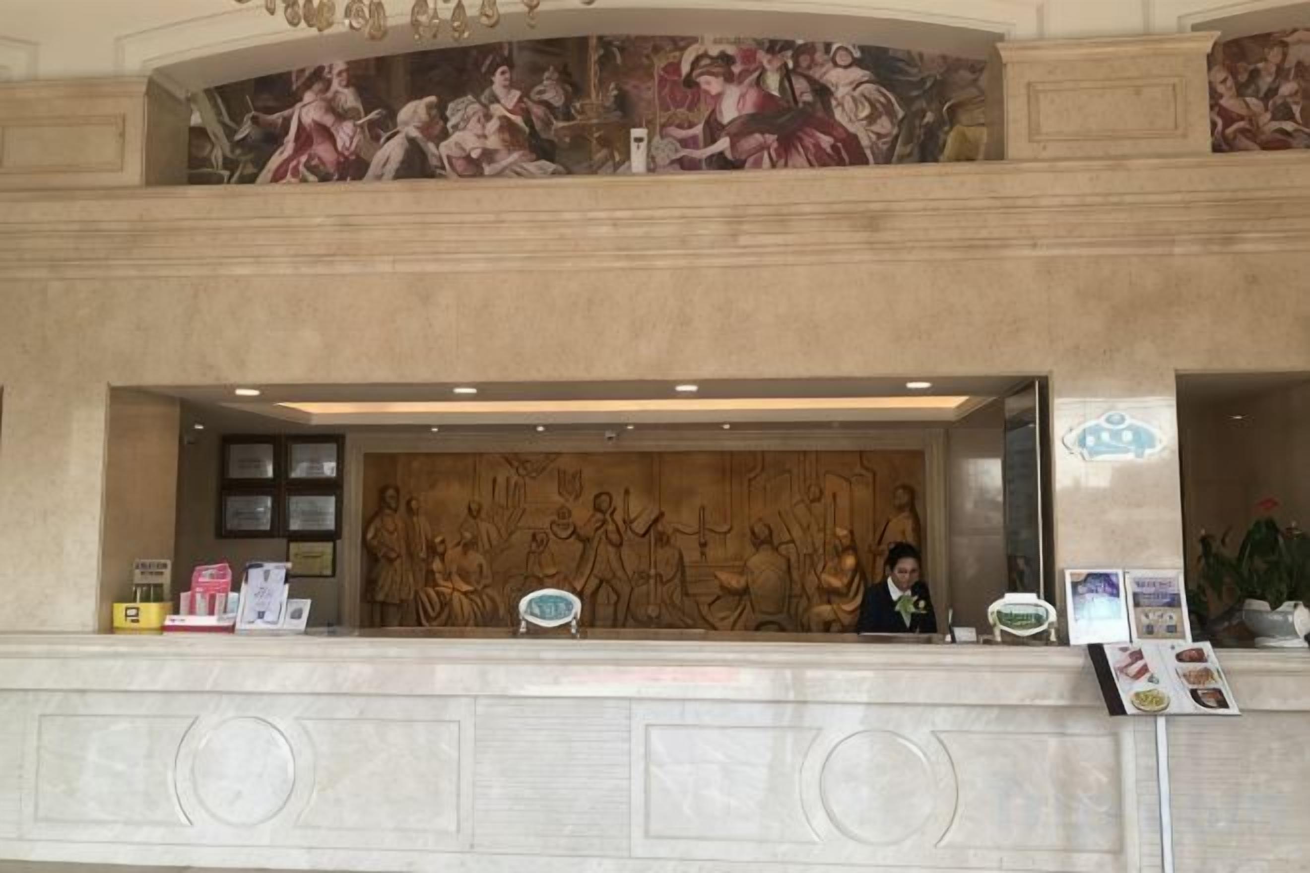 Zona de recepción de un hotel con un mostrador de mármol, una recepcionista y un mural en la pared.