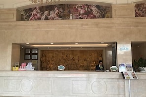 A hotel reception area with a marble counter, a receptionist, and a mural above. - Vienna Hotel (Jieyang Longwei) (Jieyang)