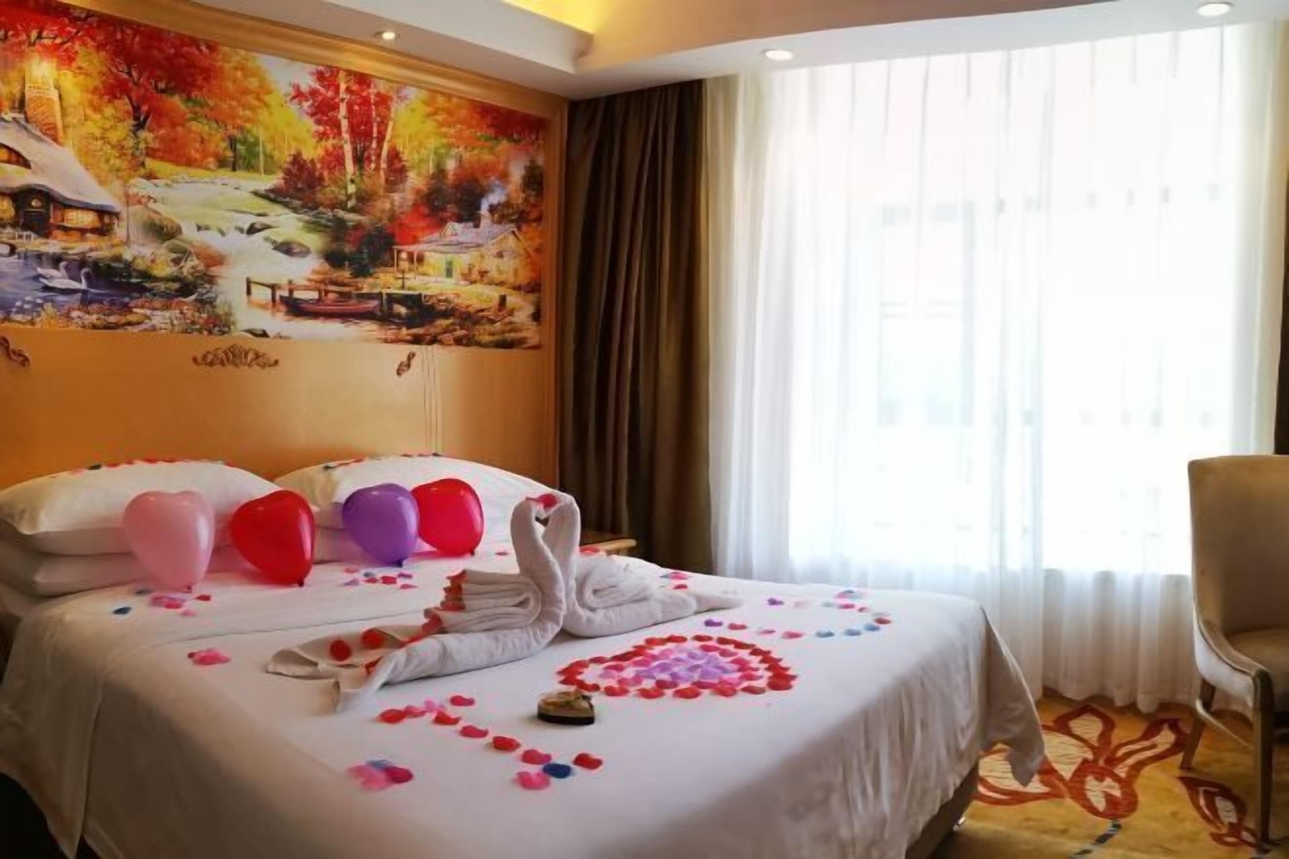 A hotel room with a bed adorned with heart-shaped pillows, a swan-shaped towel, and rose petals. A painting of a scenic landscape is on the wall, and the room has a large window with curtains.