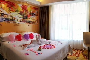 Habitación de hotel con una cama decorada con almohadas con forma de corazón, una toalla con forma de cisne y pétalos de rosa. Un cuadro de un paisaje pintoresco adorna la pared, y la habitación tiene un amplio ventanal con cortinas.